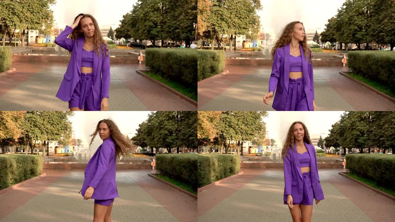 一个美丽苗条的卷发女孩正沿着城市的一条小巷走，慢莫。一个穿着紫色时尚西装的年轻女孩。