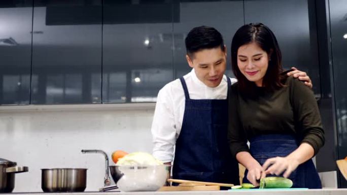 亚洲夫妇在家里的现代厨房里一起做饭做饭，妻子正在切黄瓜做蔬菜沙拉，把黄瓜喂给丈夫吃美味