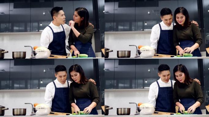 亚洲夫妇在家里的现代厨房里一起做饭做饭，妻子正在切黄瓜做蔬菜沙拉，把黄瓜喂给丈夫吃美味