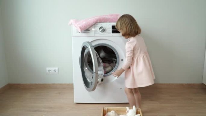 小快乐的3岁女孩站在洗衣机旁边，打开它，微笑着放柔软的玩具进行洗涤并关闭舱门
