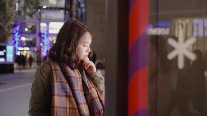 亚洲女孩女人站在商店橱窗外，在寒冷的夜晚在城市里寻找圣诞节礼物，她穿着保暖的衣服。