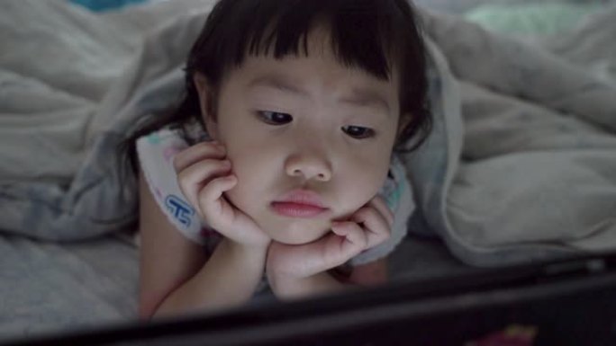 可爱的亚洲小女孩躺在毯子下晚上在卧室看数字笔记本电脑。手持镜头，真实直播
