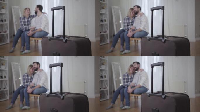 手提箱的特写镜头站在前台，幸福的年轻夫妇拥抱在后台。已婚男女旅行后见面。生活方式、婚姻、团结、幸福