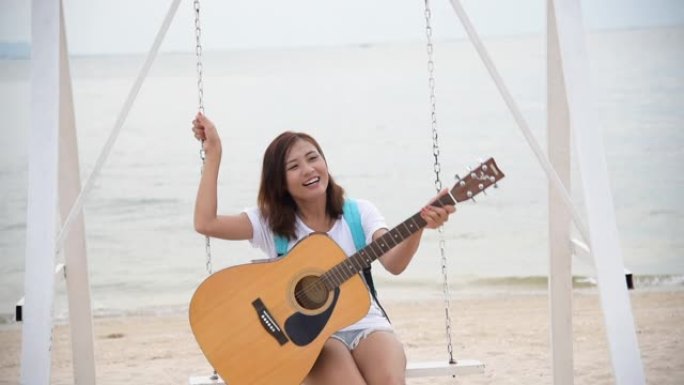 美丽的亚洲可爱女孩在海滩上玩木吉他。女人穿着休闲白色t恤，坐在热带海滩夏岛上，带着幸福的笑脸。自由旅
