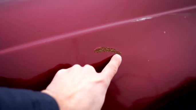 男子在汽车油漆中显示有锈迹的划痕。手指在红色汽车上刮擦