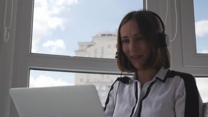 年轻成功的女性穿着严格的衣服和耳机坐在现代办公室，在与同事的视频会议上通过网络摄像头讲话