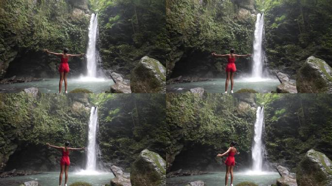 身穿红色连体衣的苗条年轻女子在风景优美的瀑布前举起双手。