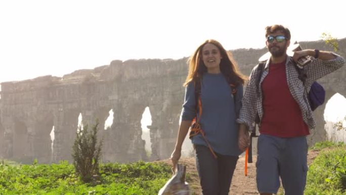 年轻夫妇背包客游客在迷雾日出时带着吉他和睡袋慢动作走向罗马公园遗址的罗马渡槽拱门