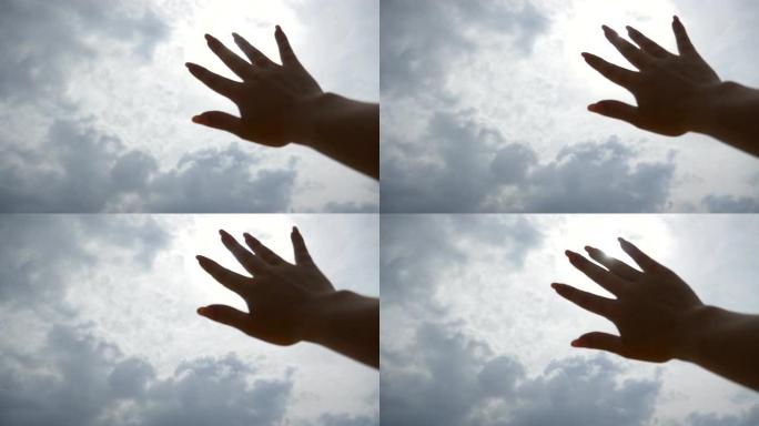 明亮的阳光穿透了年轻女子伸出手臂的手指，在多云的天空背景下。女性手的特写伸向太阳。暑假或假期的概念。