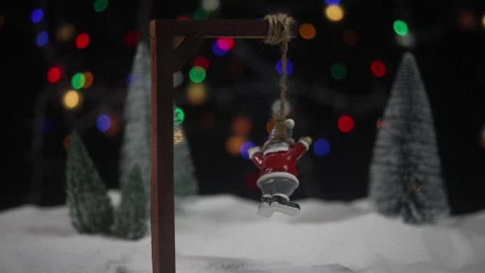 有趣的新年概念。圣诞老人雕像被倒吊在脚手架上。雪地上带有圣诞属性的创意艺术品装饰。