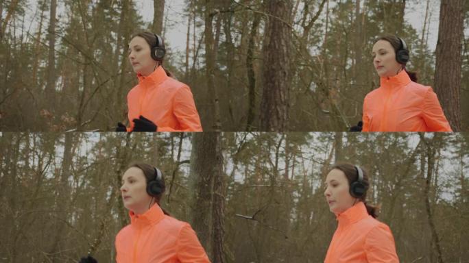 在公园慢跑的女人。穿着耳机和橙色夹克的自信专注的女运动员早上在冬季森林跑步。职业跑友刻苦训练备战半程