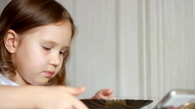 孩子用勺子吃汤，然后在智能手机上观看视频。特写镜头女婴坐在家里做饭和吃饭的桌子上