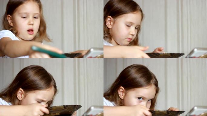 孩子用勺子吃汤，然后在智能手机上观看视频。特写镜头女婴坐在家里做饭和吃饭的桌子上