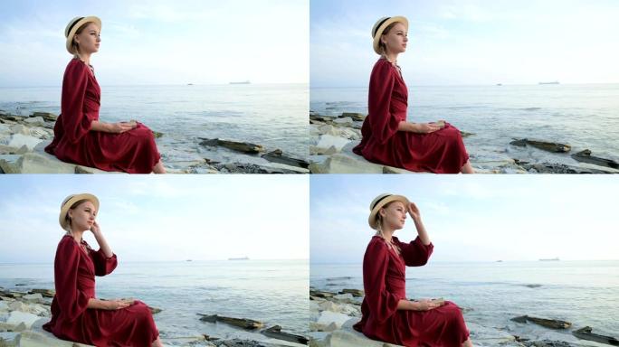 一个迷人的高加索女孩的肖像，穿着一件夏天的红色连衣裙，戴着一顶草帽，怀里抱着一本书坐在海边的石头上，