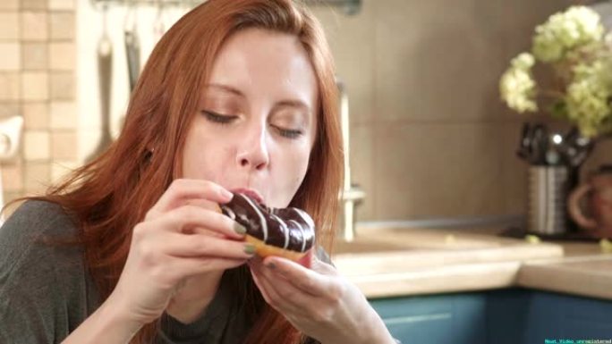 迷人的饥饿的红发女孩在周末的清晨在厨房里吃甜甜圈。不健康的饮食，高脂肪的食物，年轻时的不良生活方式