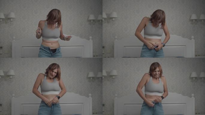 沮丧的年轻女子由于腹部脂肪而无法合上牛仔裤。女性需要减肥。卧室里慢动作的女人。