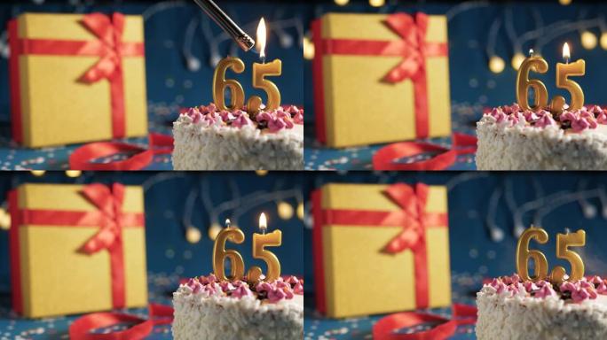 白色生日蛋糕编号65点火器燃烧的金色蜡烛，蓝色背景带灯和用红丝带绑起来的礼物黄色盒子。特写