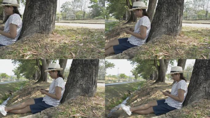 穿着便服的亚洲女性坐在树下，使用手机与社交在线。