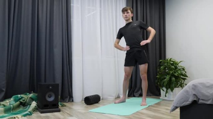 年轻的高加索男孩在室内做健身运动。穿着黑色运动服的运动男人在家做瑜伽垫有氧和康复训练。男运动员在室内