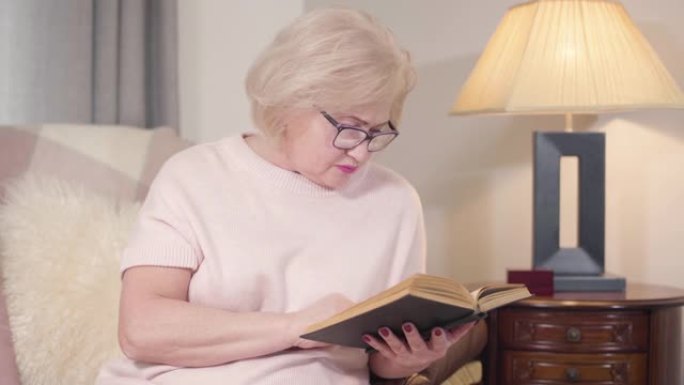 戴眼镜的白人金发女人的肖像在家看书。成熟聪明的女人晚上在室内休息。爱好，生活方式，文学