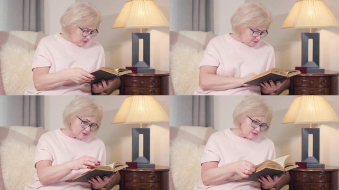 戴眼镜的白人金发女人的肖像在家看书。成熟聪明的女人晚上在室内休息。爱好，生活方式，文学