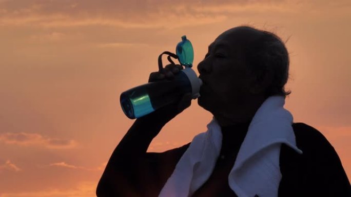 特写剪影亚洲老年人在早晨的日出运动后喝水。健康生活方式和运动理念。慢动作