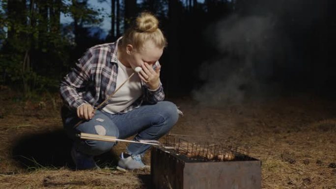 年轻女孩吃烧烤炉上油炸的木棒上的热棉花糖。晚上在森林野餐上烤棉花糖的少女。