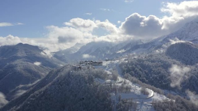 来自飞行无人机的多云天空景观上雪峰上的山村。鸟瞰雪山度假村的蜿蜒道路。福吉天空背景上的雪山和林地令人
