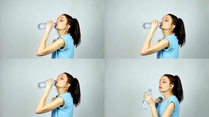 深色长发的黑发女孩在工作室的灰色背景下锻炼后，正在喝塑料透明瓶子里的纯净水
