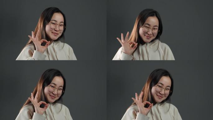 年轻的亚洲女子在灰色背景上做OK标志。赢家。成功。积极的韩国女孩对镜头微笑。肢体语言。