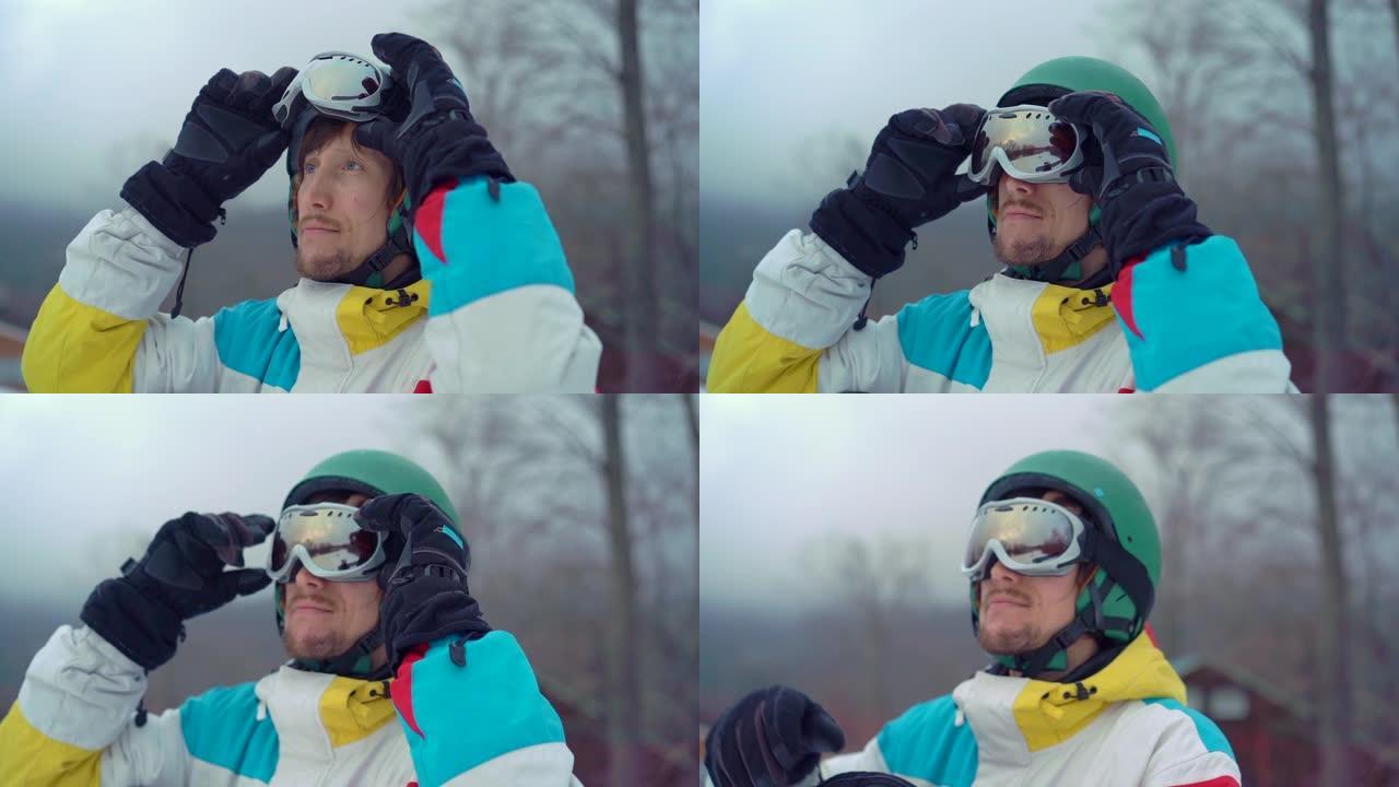 在山区度假胜地，一个戴着头盔和滑雪板的年轻人的特写镜头。他穿上雪地谷歌。寒假概念。慢动作镜头