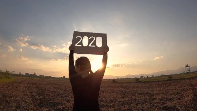 女人的剪影拿着回收纸板，2020文本在日落天空背景和阳光透过文本。新年和庆祝活动的概念。