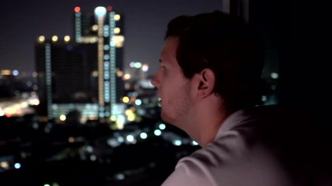 悲伤的年轻人在夜晚以城市天际线为背景的高层建筑的窗外