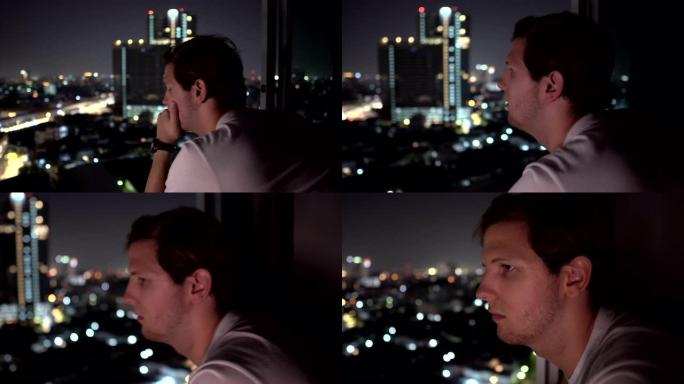 悲伤的年轻人在夜晚以城市天际线为背景的高层建筑的窗外