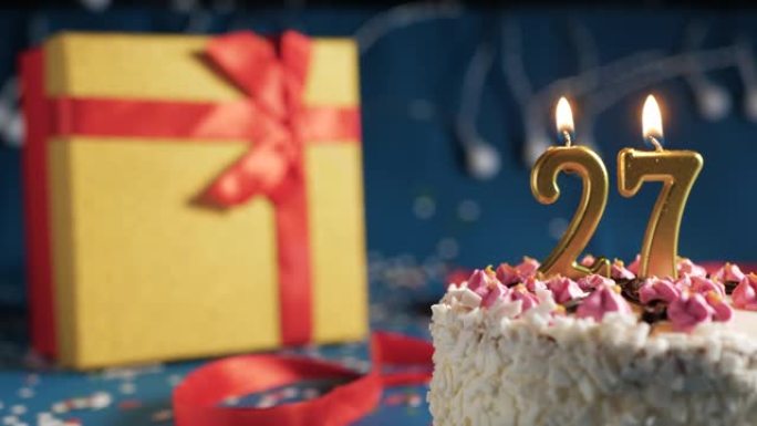 白色生日蛋糕27号点火器燃烧的金色蜡烛，蓝色背景带灯和用红丝带绑起来的礼物黄色盒子。特写
