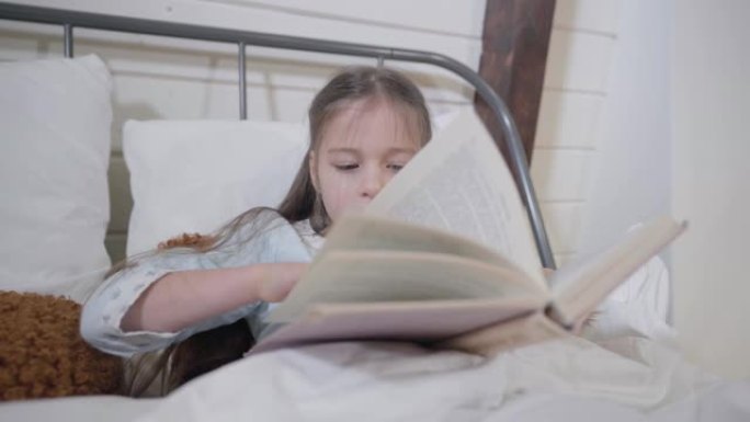 漂亮的高加索女孩躺在床上看书的肖像。聪明的孩子晚上睡觉。就寝时间，爱好，休闲。