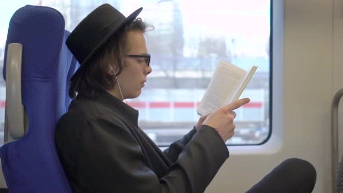 年轻英俊的时尚男子戴着黑帽子和眼镜坐在公共交通工具上看书，千禧一代的时髦学生喜欢在地铁里读书