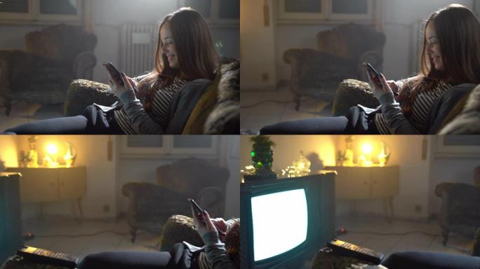 微笑的女孩坐在黄昏房间的复古扶手椅上并使用手机的特写镜头。快乐的女人拿着智能手机，同时看电视。现代电