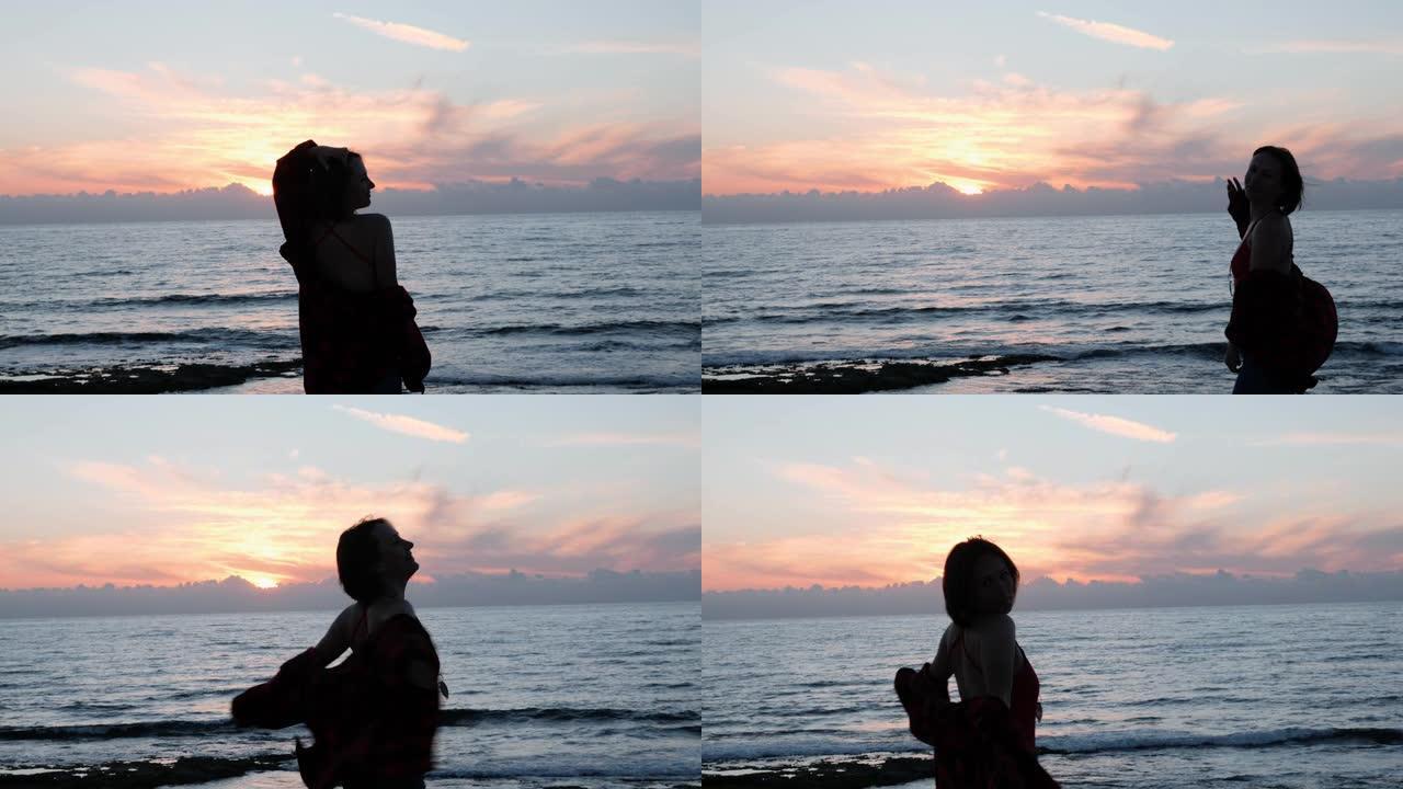 日落时分，性感的黑发女孩为相机摆姿势。年轻女子在岩石海岸微笑和旋转的剪影。黄昏时，穿着红色泳衣和牛仔