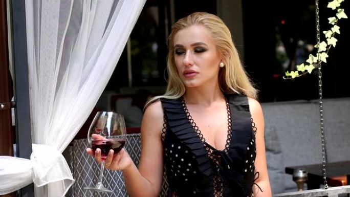 穿着黑色连衣裙的女孩坐在夏季街头咖啡馆里喝红酒。