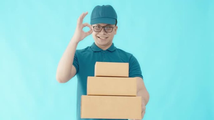 亚洲快乐送货员戴着一顶帽子，穿着一件蓝色衬衫，拿着纸质包装箱，孤立在蓝色背景上，面带微笑。邮政递送服