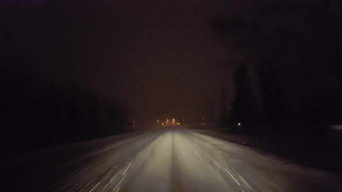 夜间在雪地暴风雪中行驶，接近乡村道路上的红绿灯。驾驶员的观点POV在冬季晚上朝着城市的灯光下雪和雪花