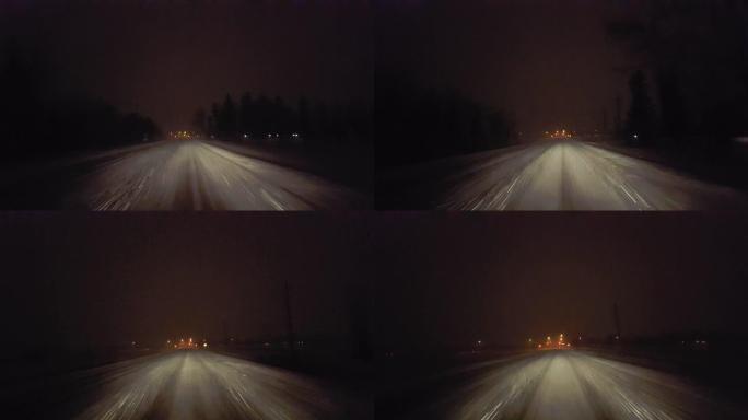 夜间在雪地暴风雪中行驶，接近乡村道路上的红绿灯。驾驶员的观点POV在冬季晚上朝着城市的灯光下雪和雪花