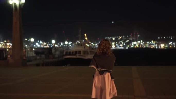 卷曲的女人后视图在晚上，强风，在夜市的灯光背景下，去船上。