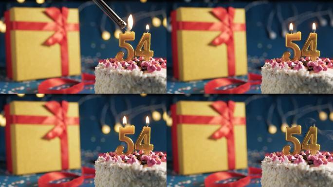 白色生日蛋糕编号54点灯燃烧的金色蜡烛，带灯的蓝色背景和用红丝带绑起来的礼物黄色盒子。特写