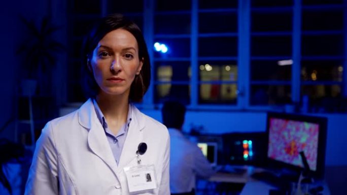 自信的女科学家在白色实验室外套上拿着身份证的中等镜头在实验室里摆姿势看着相机，她的男同事在后台的笔记