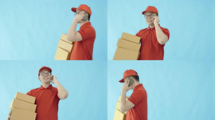 亚洲快乐送货员戴着帽子和红色衬衫，使用智能手机，携带纸质包装箱，孤立在蓝色背景上，面带微笑。邮政递送
