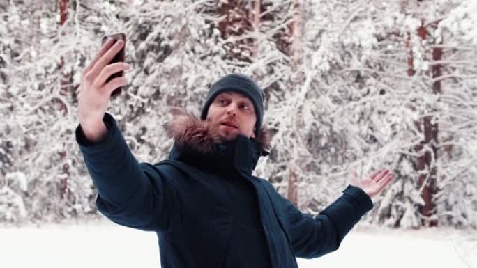 一位帅气的视频博主的肖像旅游男子在白雪皑皑的冬林中使用手机，自拍，视频会议，视频博主
