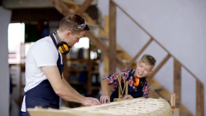 中年木匠建造大型木船模型的侧视图，他好奇的小儿子站在木工车间的工作台上探索过程并提问