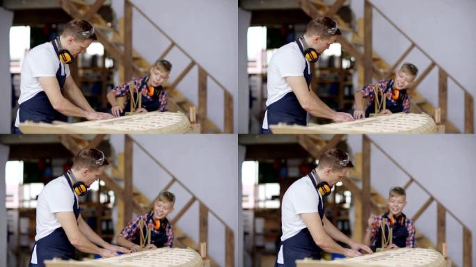 中年木匠建造大型木船模型的侧视图，他好奇的小儿子站在木工车间的工作台上探索过程并提问